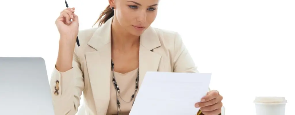 Obrazek wyróżniający do artykułu - podanie o podwyżkę. Na zdjęciu znajduje się kobieta w jasnym, eleganckim stroju, która siedzi przy biurku i uważnie czyta dokument. W prawej ręce trzyma kartkę papieru, a w lewej ma uniesiony długopis, jakby była gotowa do zanotowania czegoś. Na biurku, po jej lewej stronie, znajduje się laptop, a po prawej stronie kubek z napojem. Tło zdjęcia jest jasne, co sugeruje, że znajduje się w dobrze oświetlonym pomieszczeniu. Kobieta wygląda na skoncentrowaną na czytanym dokumencie.