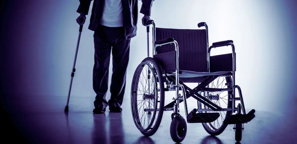 Na obrazie widać pusty wózek inwalidzki ustawiony na pierwszym planie, symbolizujący różne stopnie niepełnosprawności. W tle widać osobę wspierającą się na kulach, co podkreśla różnorodność doświadczeń osób z niepełnosprawnością. Tło jest lekko rozmyte, co nadaje zdjęciu melancholijny nastrój, wywołując refleksję nad codziennymi wyzwaniami i siłą ludzi mierzących się z różnymi ograniczeniami.