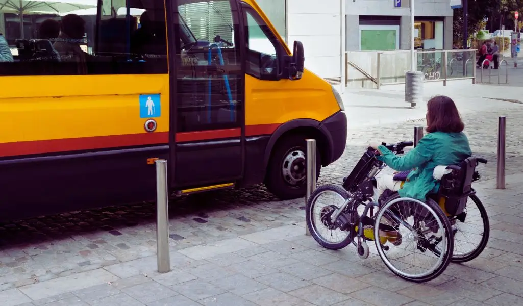 Jakie możliwości ma osoba na wózku inwalidzkim w ruchu pieszym i podróży? 1 Na zdjęciu osoba na wózku inwalidzkim, zbliża się do żółtego busa oznaczonego symbolem dostępności dla osób z niepełnosprawnościami. Pojazd stoi na miejskim chodniku, a między nim a osobą na wózku znajdują się metalowe słupki zabezpieczające. Scena może budzić różne emocje, takie jak poczucie niezależności, ale też pewne wyzwanie związane z dostępnością przestrzeni miejskiej. Obrazek pokazuje codzienną sytuację, z którą osoby z ograniczoną mobilnością mogą się zmierzyć, podkreślając znaczenie przystosowania infrastruktury do ich potrzeb.