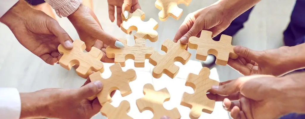 Na zdjęciu widać kilka rąk różnych osób trzymających drewniane elementy układanki – puzzle. Każda z osób trzyma jeden kawałek, a wszystkie elementy są skierowane do środka, jakby miały za chwilę stworzyć jedną spójną całość. Obrazek symbolizuje współpracę, wzajemne wsparcie i dążenie do wspólnego celu. To wizualna metafora na cechy dobrego pracownika – takie jak umiejętność pracy zespołowej, zaangażowanie, komunikatywność i odpowiedzialność. Zdjęcie budzi pozytywne emocje: poczucie jedności, wspólnoty i harmonii w działaniu.