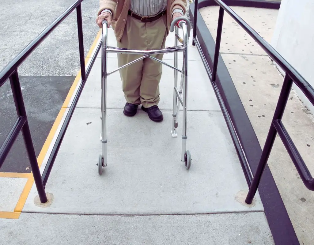 Starsza osoba korzystająca z chodzika wchodzi na podjazd dla niepełnosprawnych, otoczony czarnymi poręczami po obu stronach. Betonowa nawierzchnia jest równa, a przestrzeń zapewnia bezpieczeństwo i samodzielność. Obraz oddaje poczucie niezależności, spokoju i ulgi – pokazuje, jak ważna jest dostępność przestrzeni publicznej dla osób z ograniczoną mobilnością.